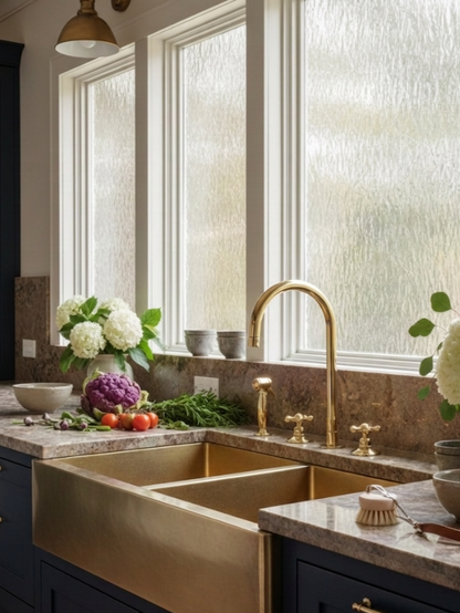 Unlacquered brass three-hole bridge kitchen faucet with side sprayer installed with a brass farmhouse sink.