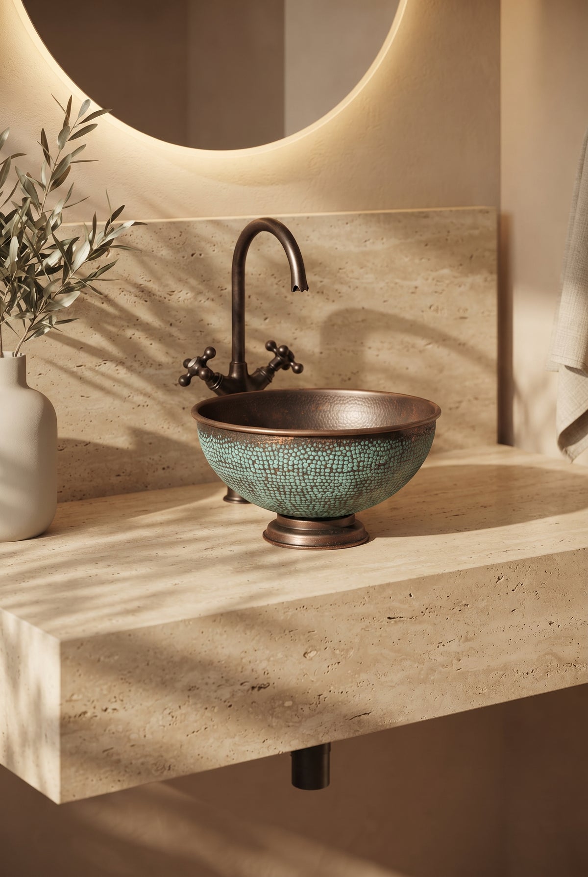 Aged copper vessel sink with turquoise patina and a pedestal base on a beige stone vanity beneath a curved faucet and round mirror.