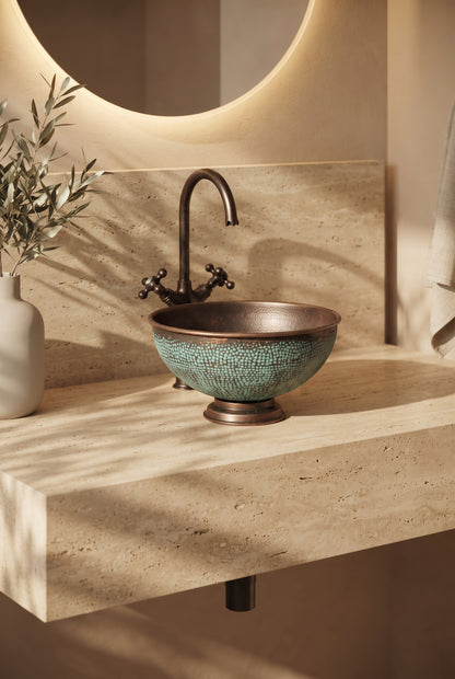 Aged copper vessel sink with turquoise patina and a pedestal base on a beige stone vanity beneath a curved faucet and round mirror.