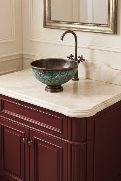 Round copper vessel sink with turquoise patina on a cream countertop above burgundy cabinetry, paired with a dark curved faucet.