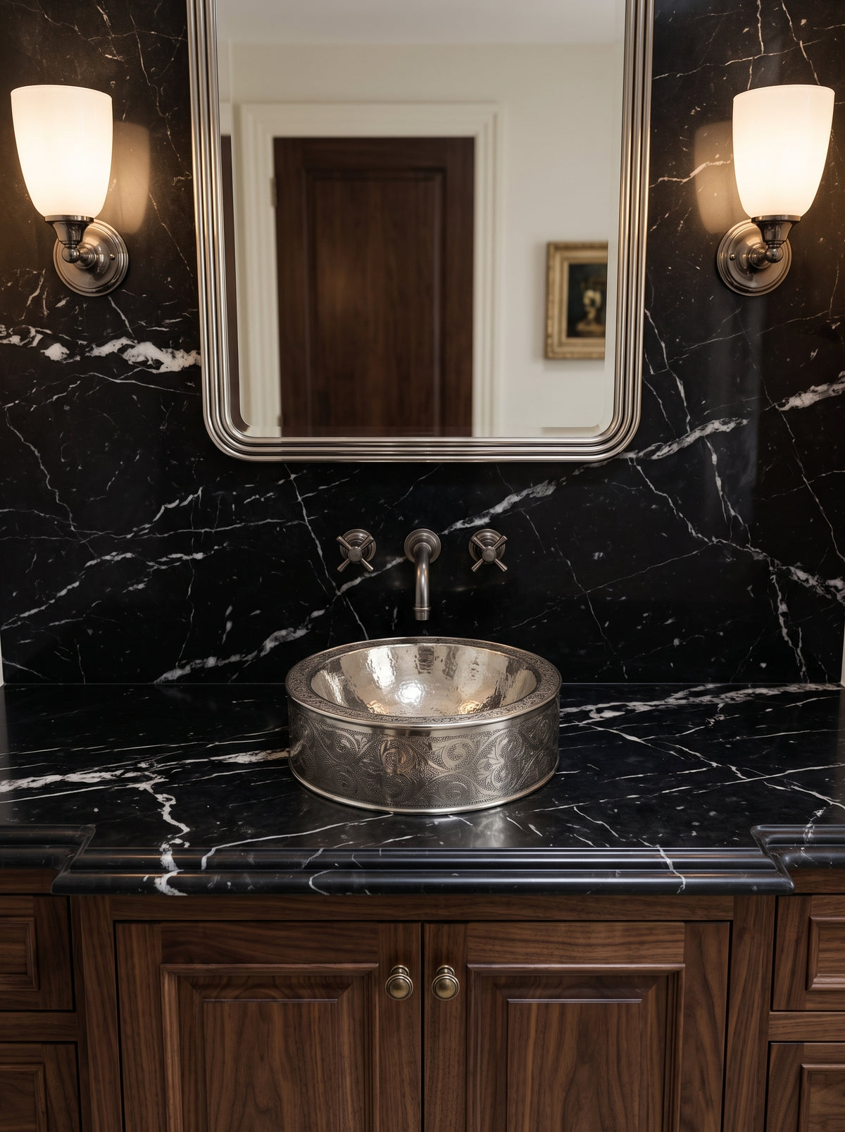 Engraved silver vessel sink on a black marble vanity with matching wall-mounted faucet, rectangular mirror, and illuminated sconces.