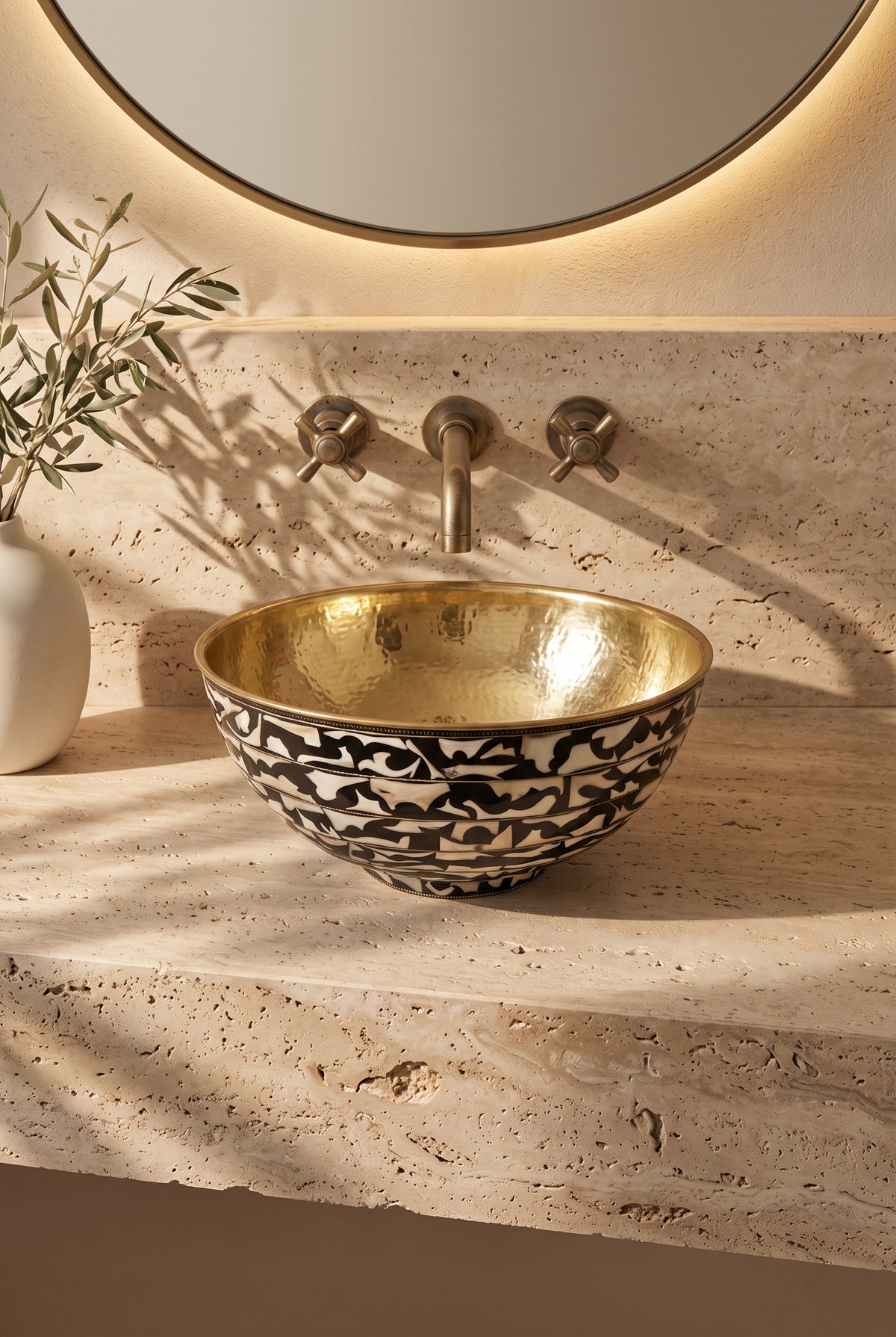 Round vessel sink with a hammered brass interior and black-and-white floral bone inlay exterior on beige stone beneath a wall-mounted faucet.