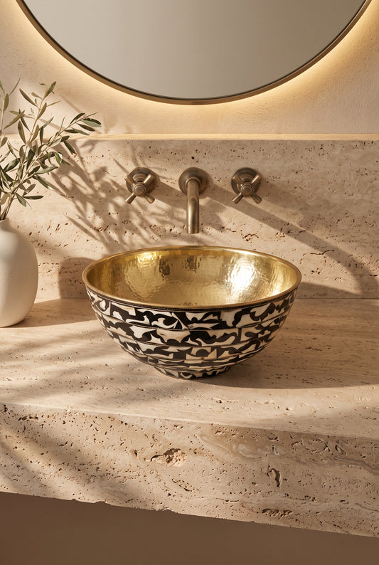 Round vessel sink with a hammered brass interior and black-and-white floral bone inlay exterior on beige stone beneath a wall-mounted faucet.