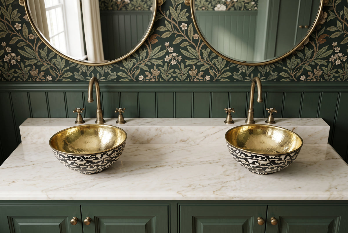 Two brass vessel sinks with black-and-white bone inlay exteriors on a marble countertop above green cabinets, beneath round mirrors and botanical wallpaper.