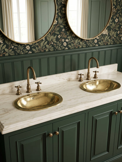Two oval hammered brass sinks in a cream marble double vanity with brass faucets, green cabinets, floral wallpaper, and oval mirrors.