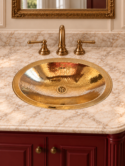 Oval hammered brass sink in a cream marble countertop with a brass faucet, burgundy vanity, and ornate gold-framed mirror.
