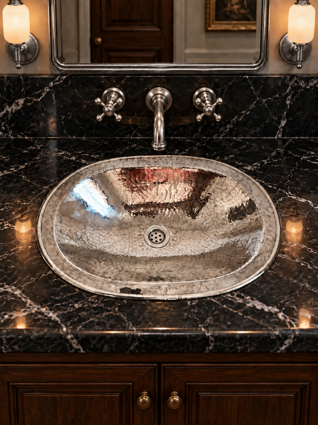 Oval hammered silver sink in a black marble countertop with a matching wall-mounted faucet, silver-framed mirror, and illuminated sconces.