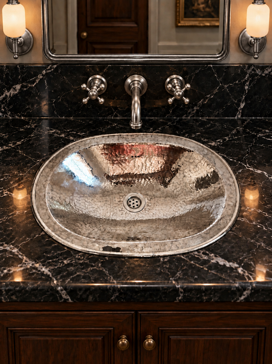 Oval hammered silver sink in a black marble countertop with a matching wall-mounted faucet, silver-framed mirror, and illuminated sconces.
