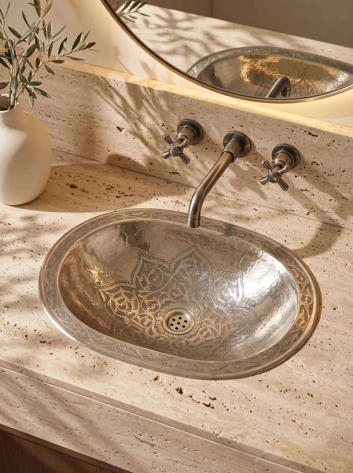Oval silver sink with engraved floral motifs and decorative rim in a travertine vanity beneath a wall-mounted faucet and round mirror.