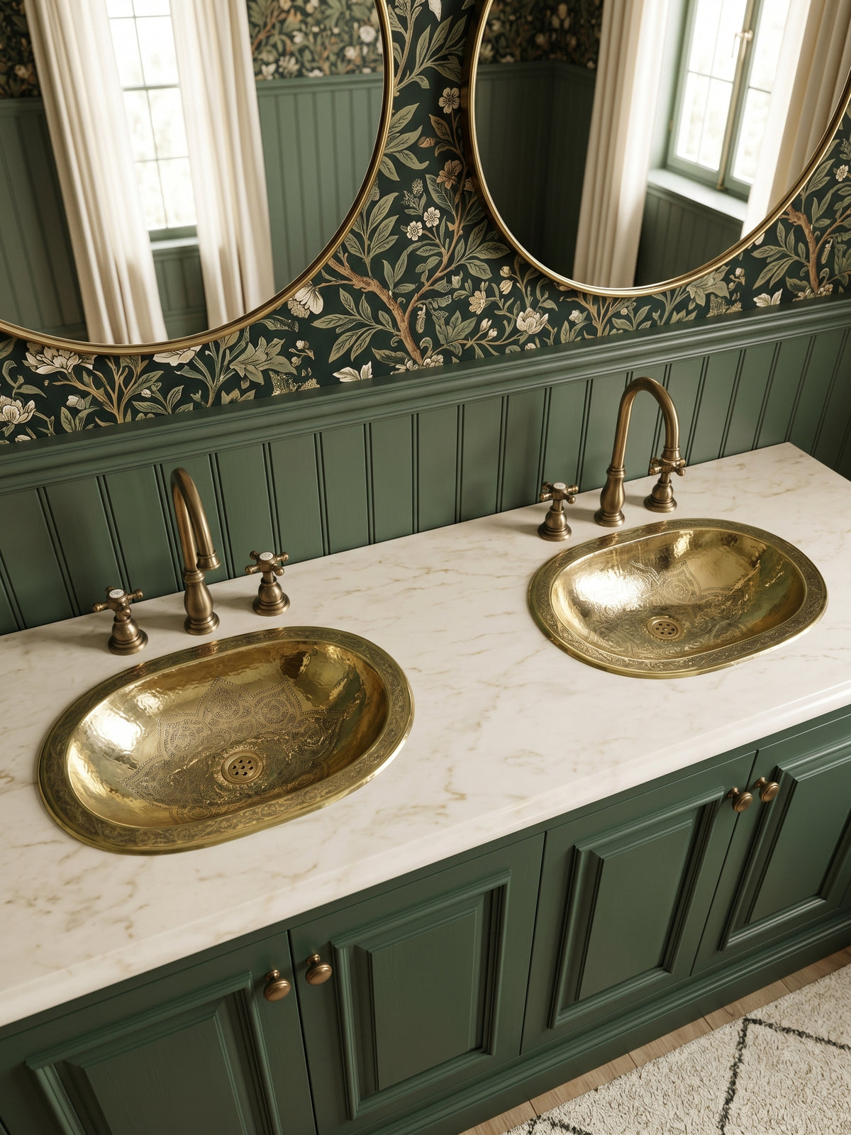 Two engraved oval brass sinks in a white marble double vanity with green cabinets, brass faucets, floral wallpaper, and oval mirrors.