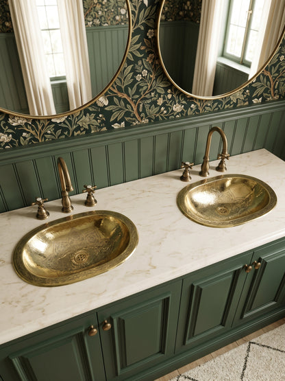 Two engraved oval brass sinks in a white marble double vanity with green cabinets, brass faucets, floral wallpaper, and oval mirrors.
