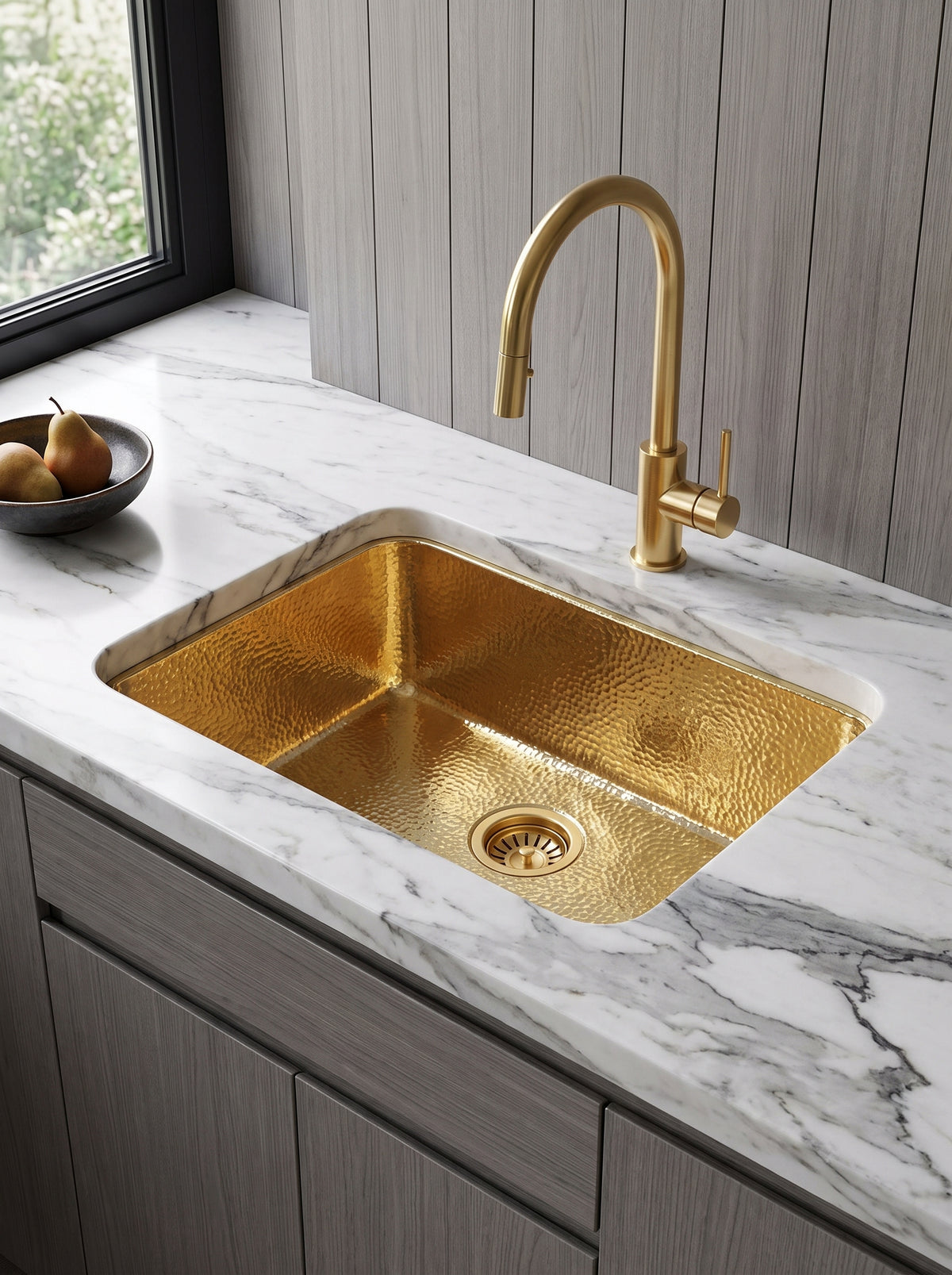 Rectangular hammered brass undermount sink with matching gooseneck faucet in white marble, above gray wood cabinets beside a window.