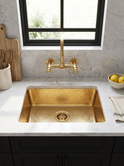 Hammered brass undermount sink beneath a wall-mounted brass faucet and black-framed window, surrounded by white marble above black cabinets.