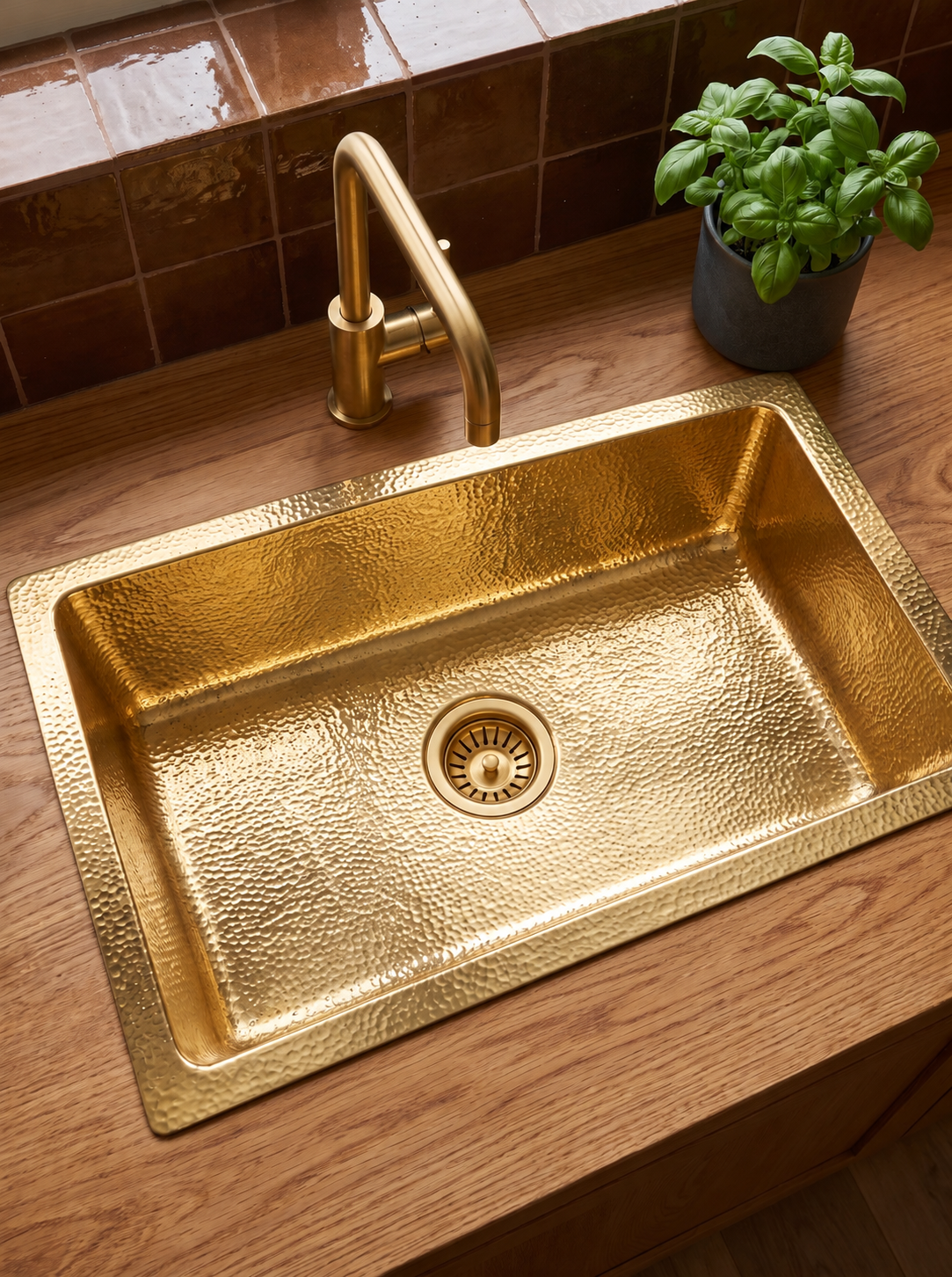 Rectangular hammered brass drop-in sink with matching faucet on an oak countertop, beside a basil plant and brown tile backsplash.