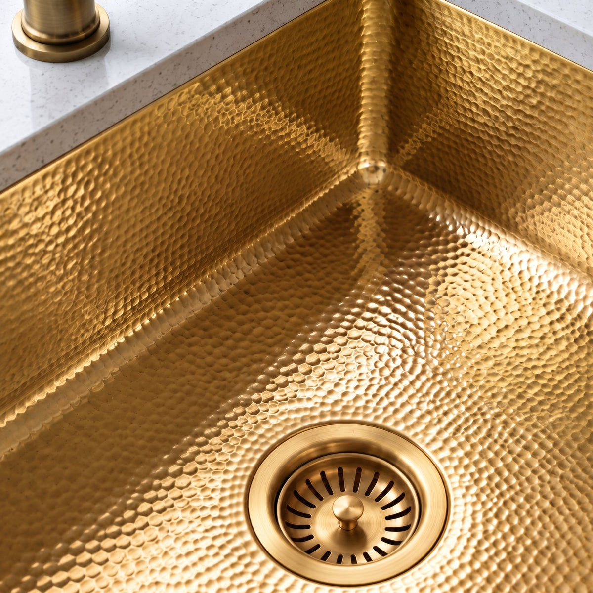 Close-up of a hammered brass sink showing its textured basin, rounded interior corner, and matching basket-strainer drain.