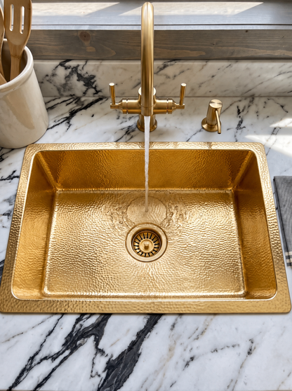 Hammered brass drop-in sink with water flowing from a matching bridge faucet, set into white marble with bold dark veining.