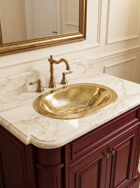 Angled view of an oval engraved brass sink in a cream marble countertop with a brass faucet, burgundy vanity, and gold-framed mirror.