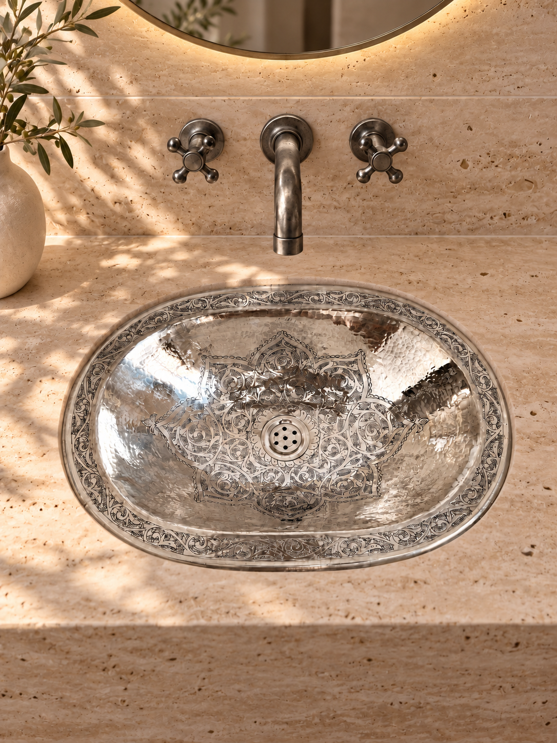 Oval engraved silver sink in a beige travertine vanity beneath a wall-mounted silver faucet and round backlit mirror.