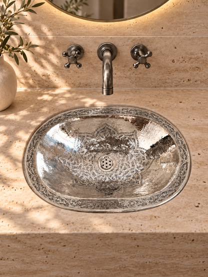 Oval engraved silver sink in a beige travertine vanity beneath a wall-mounted silver faucet and round backlit mirror.