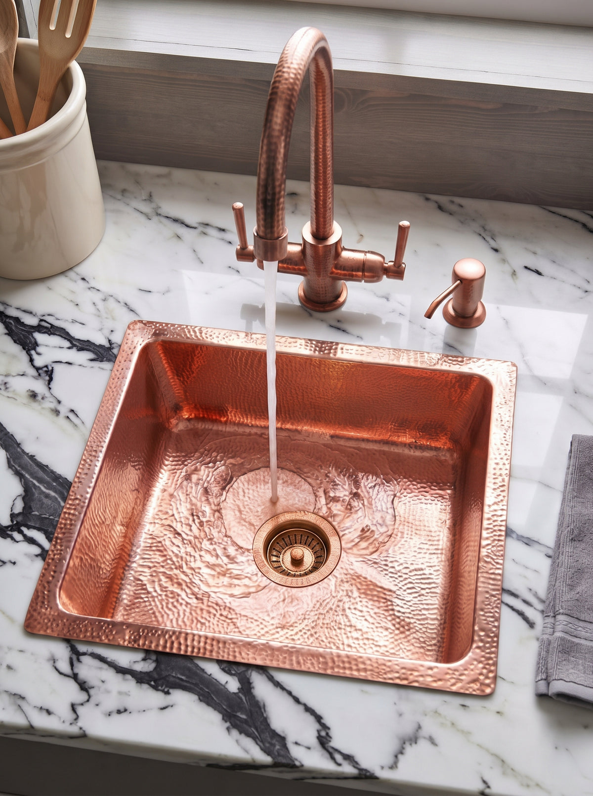 Hammered copper drop-in sink with running water from a matching bridge faucet, set into white marble with bold dark veining.