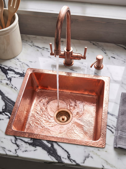 Hammered copper drop-in sink with running water from a matching bridge faucet, set into white marble with bold dark veining.