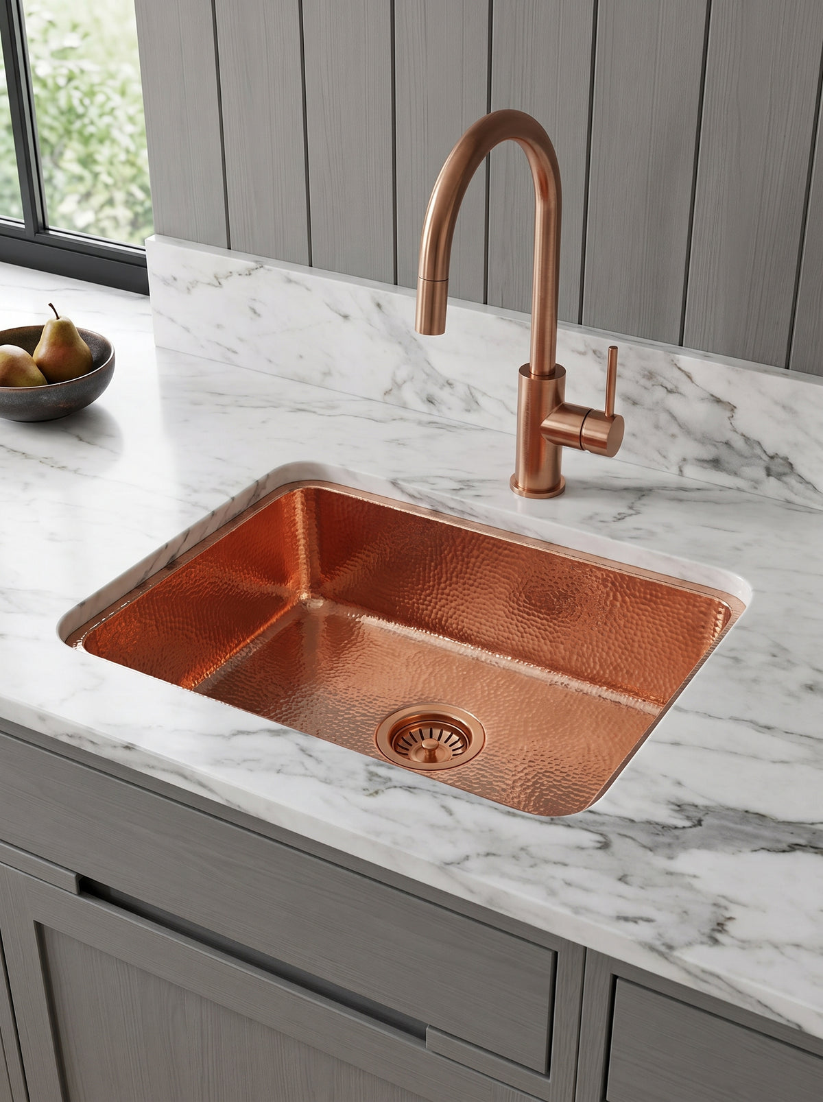 Rectangular hammered copper undermount sink with matching gooseneck faucet in white marble, above gray cabinetry beside a window.