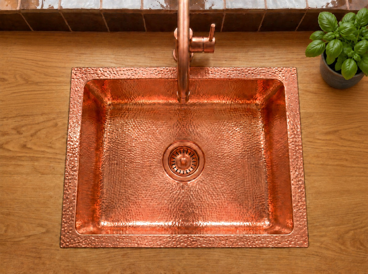 Overhead view of a hammered copper drop-in sink with matching faucet on an oak countertop, beside a basil plant and brown tile backsplash.