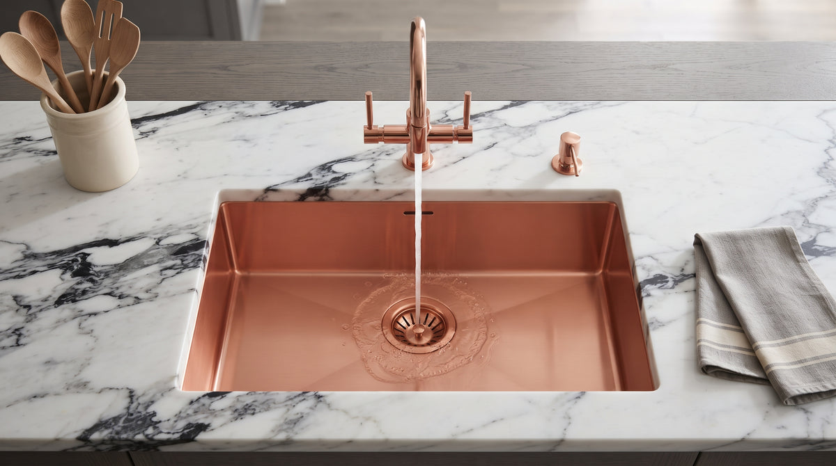 Rectangular copper kitchen sink with running water from a matching faucet, set into boldly veined white marble beside wooden utensils and a folded towel.