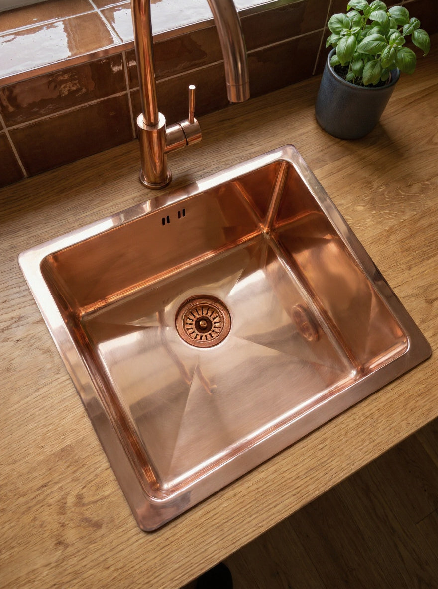 Polished copper drop-in kitchen sink with matching faucet on an oak countertop, beside a basil plant and brown tile backsplash.