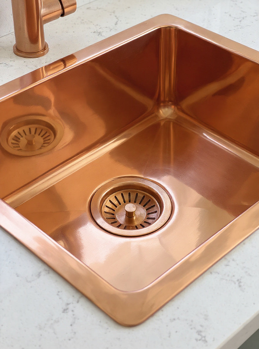 Close-up of a smooth copper sink interior and matching basket-strainer drain, with its rim resting on a pale speckled countertop.