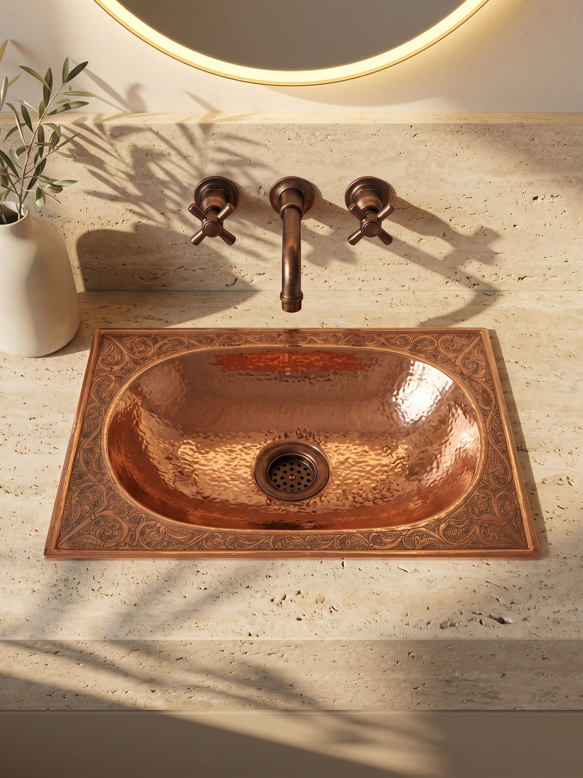 Oval copper drop-in sink with a hammered basin and engraved rectangular rim in travertine beneath a copper wall-mounted faucet and backlit mirror.