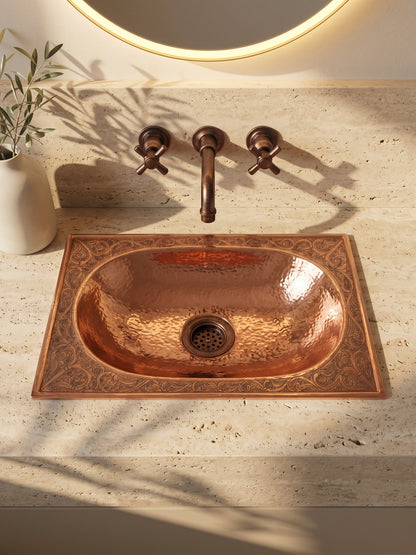 Oval copper drop-in sink with a hammered basin and engraved rectangular rim in travertine beneath a copper wall-mounted faucet and backlit mirror.