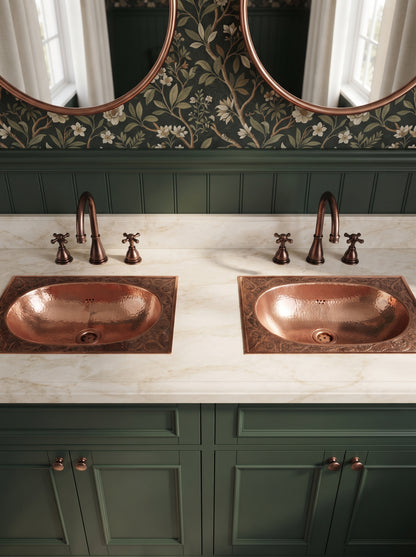 Two hammered oval copper drop-in sinks with engraved rectangular rims in a cream marble double vanity with green cabinets, copper faucets, and floral wallpaper.