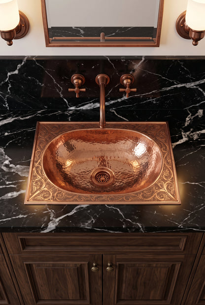 Elevated view of an oval copper drop-in sink with an engraved rectangular rim in a black marble vanity with copper faucet and wood cabinets.