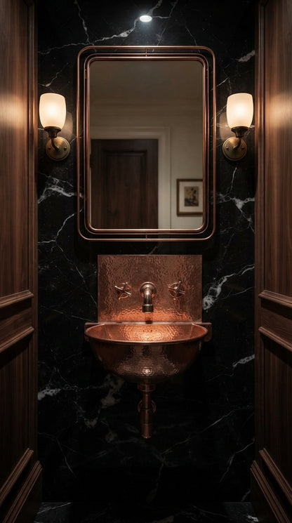 Copper wall-mounted sink with matching faucet and exposed drainpipe against black veined marble, beneath a rectangular mirror flanked by sconces.