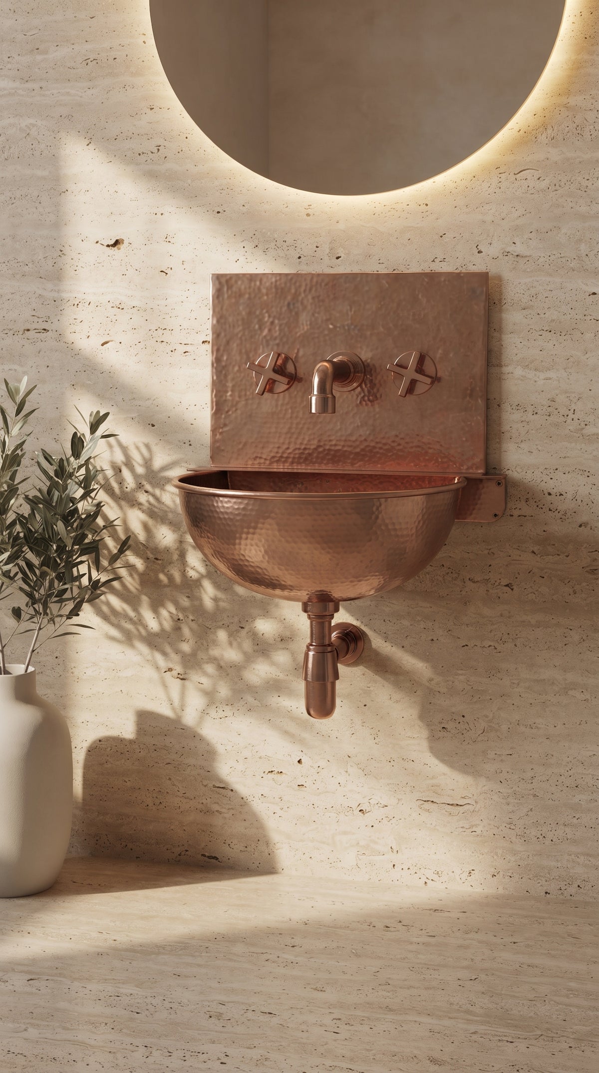 Copper wall-mounted basin with matching backsplash and faucet against beige stone, beneath a round mirror beside a vase of leafy branches.