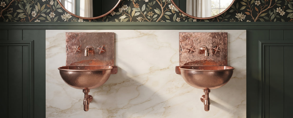 Two copper wall-mounted sinks with matching faucets and exposed plumbing against white marble, surrounded by dark green paneling and floral wallpaper.