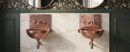 Two copper wall-mounted sinks with matching faucets and exposed plumbing against white marble, surrounded by dark green paneling and floral wallpaper.