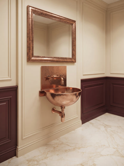 Copper wall-mounted sink with rectangular backsplash and exposed drainpipe beneath a copper-framed mirror in a cream and burgundy paneled bathroom.