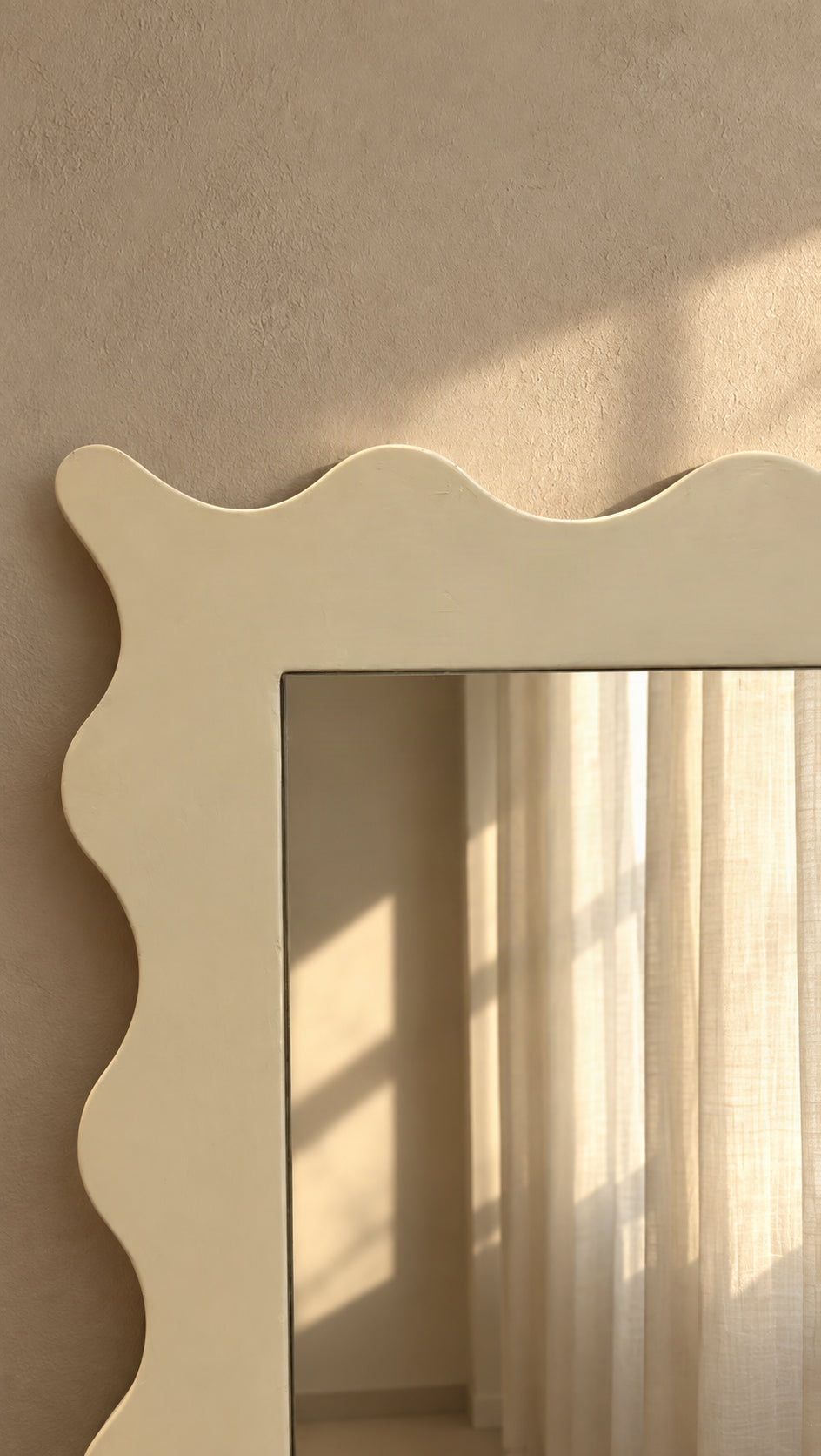 A cream-colored rectangular mirror with a decorative wavy scalloped frame, mounted on a textured beige wall. The mirror reflects soft sunlight and white curtains.
