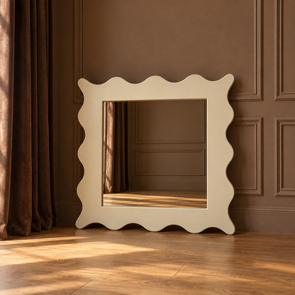 A large square wall mirror with a wavy scalloped frame in a warm cream color, leaning against a brown paneled wall on a wooden floor.