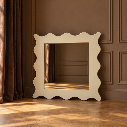A large square wall mirror with a wavy scalloped frame in a warm cream color, leaning against a brown paneled wall on a wooden floor.