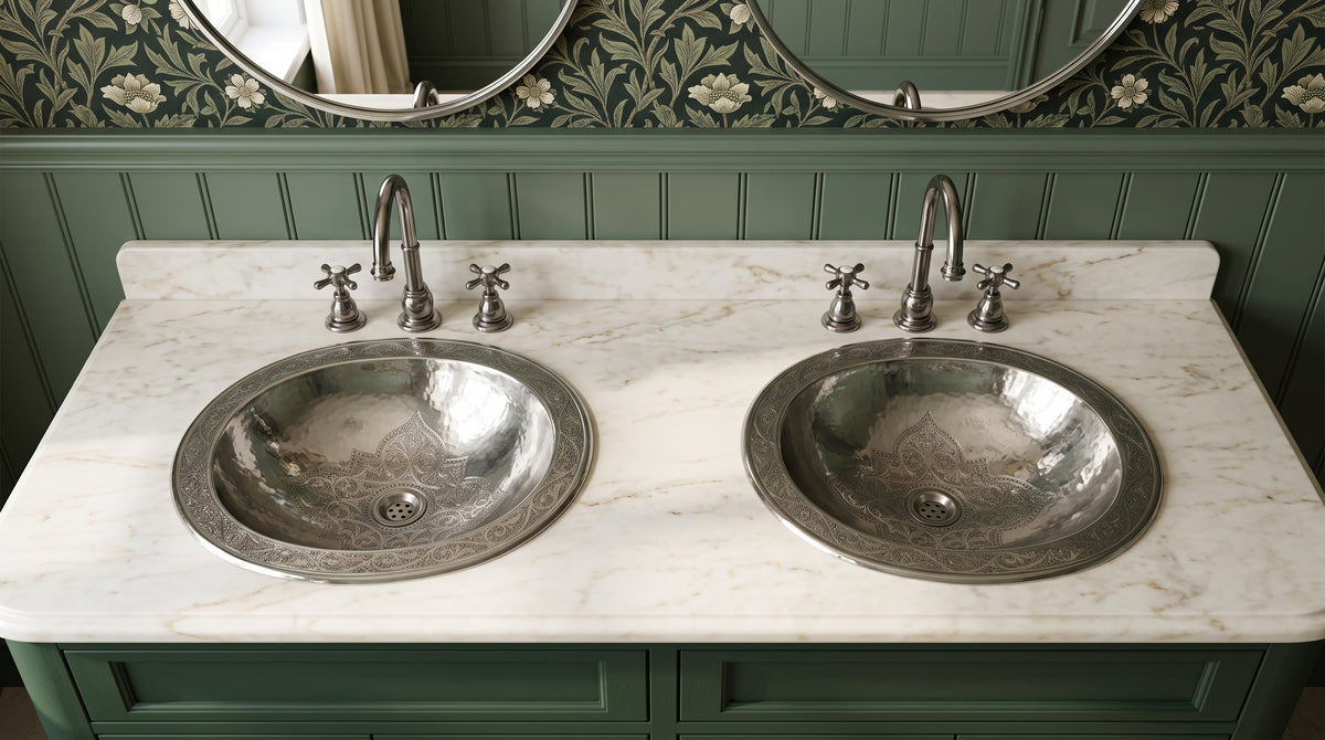 Two engraved silver drop-in sinks in a white marble double vanity with matching faucets, green cabinets, floral wallpaper, and oval mirrors.
