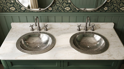 Two engraved silver drop-in sinks in a white marble double vanity with matching faucets, green cabinets, floral wallpaper, and oval mirrors.