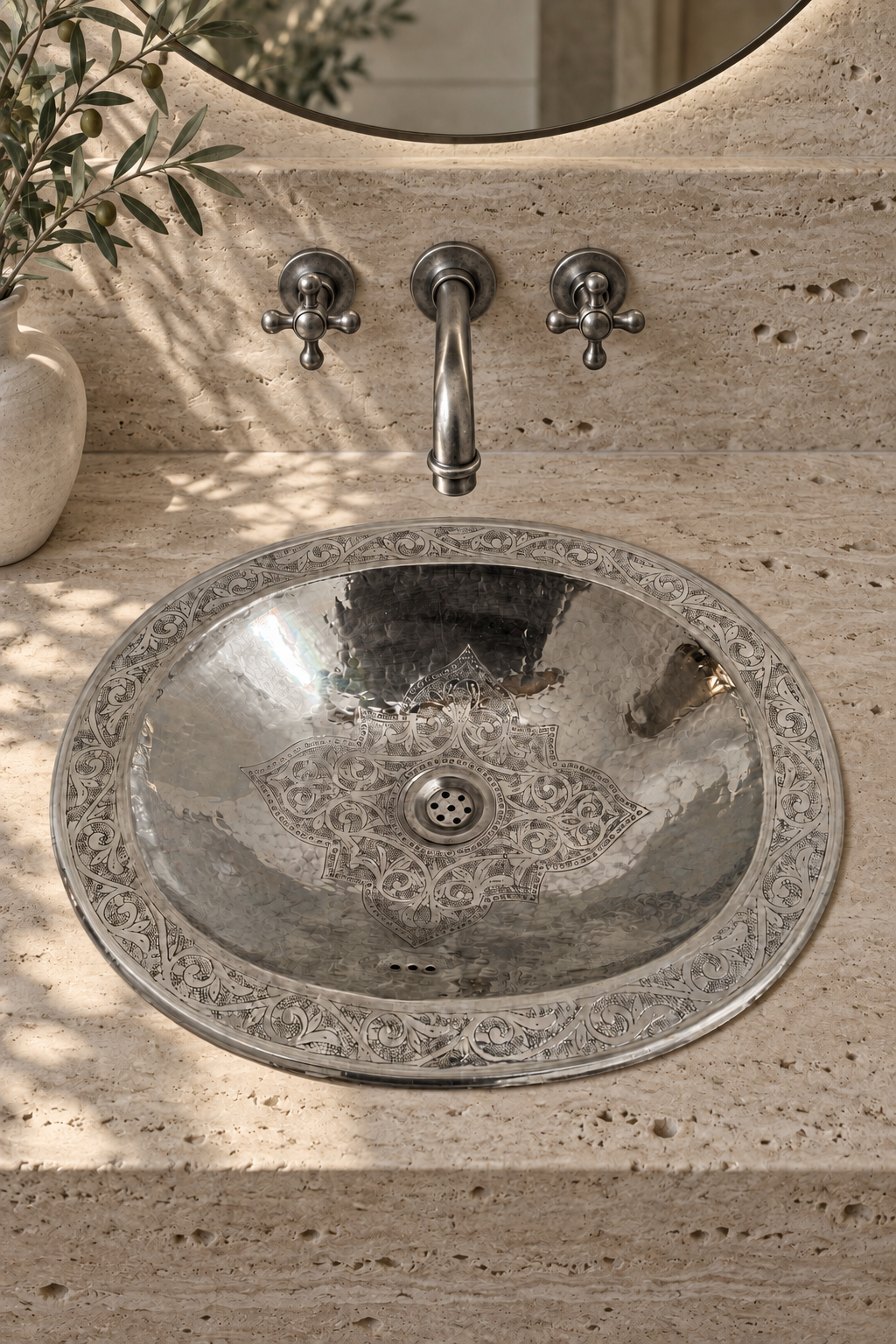 Round engraved silver sink in a beige travertine vanity beneath a wall-mounted silver faucet, beside olive branches.