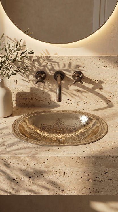 Engraved brass drop-in sink in a beige travertine vanity beneath a wall-mounted brass faucet and round backlit mirror, beside olive branches.