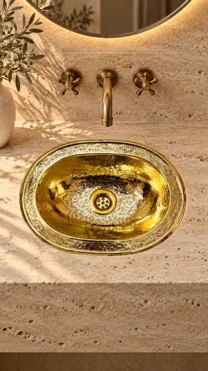 Oval brass sink with intricate engraved patterns in a beige travertine vanity beneath a wall-mounted brass faucet and round mirror.