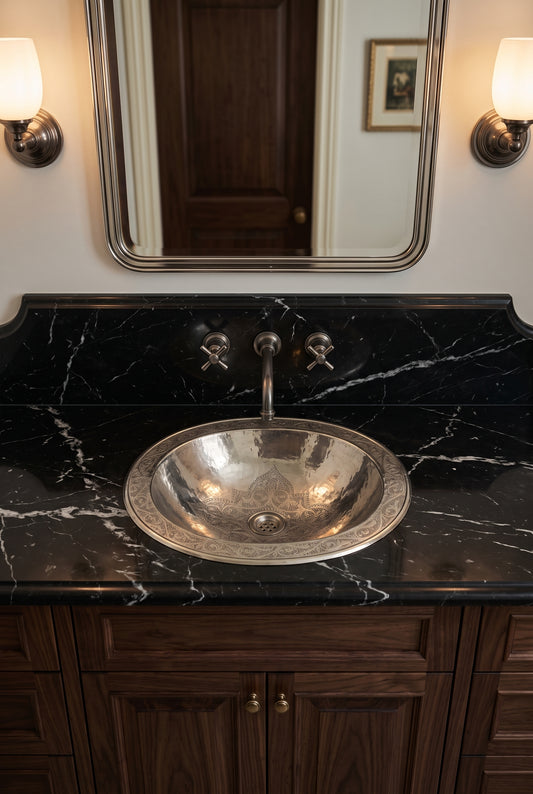 Engraved silver drop-in sink in a black marble vanity beneath a matching wall-mounted faucet, rectangular mirror, and sconces.