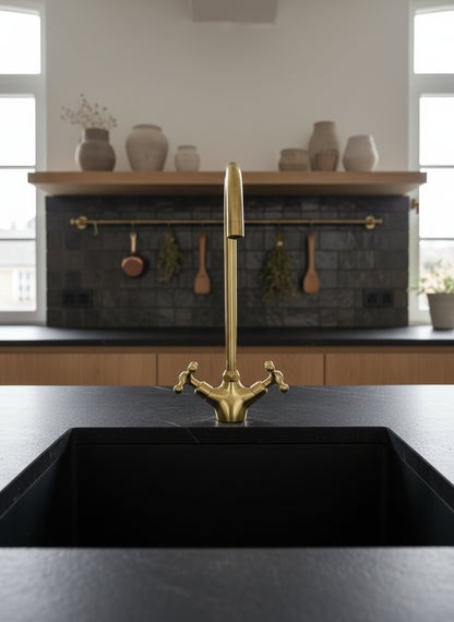 Front view of a handmade unlacquered brass bridge kitchen faucet with dual cross handles mounted above a black undermount sink.