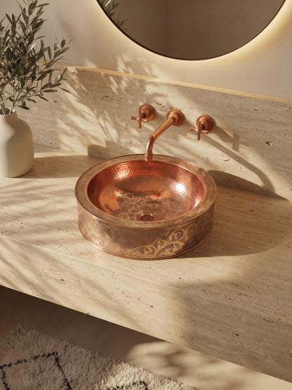 Round engraved copper vessel sink on a travertine vanity beneath a matching wall-mounted faucet and round backlit mirror.